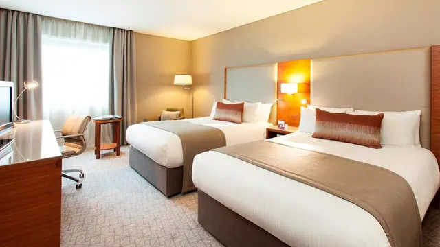 A hotel bedroom with two double beds, a desk and chair, a television, and an armchair beside a window with sheer curtains.