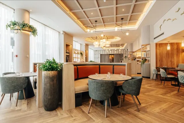 A restaurant dining room with a curved orange banquette, round tables with upholstered chairs, potted plants and sheer curtains at tall windows.