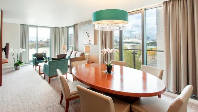 A suite living and dining area with an oval wooden table, teal armchairs and floor-to-ceiling windows looking out towards the O2 arena.