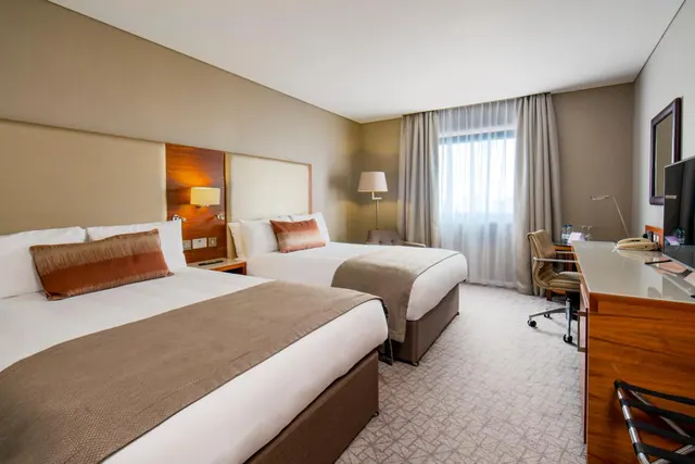 A hotel bedroom with two double beds, a desk with a chair, a wall-mounted television and a window with sheer and blackout curtains.