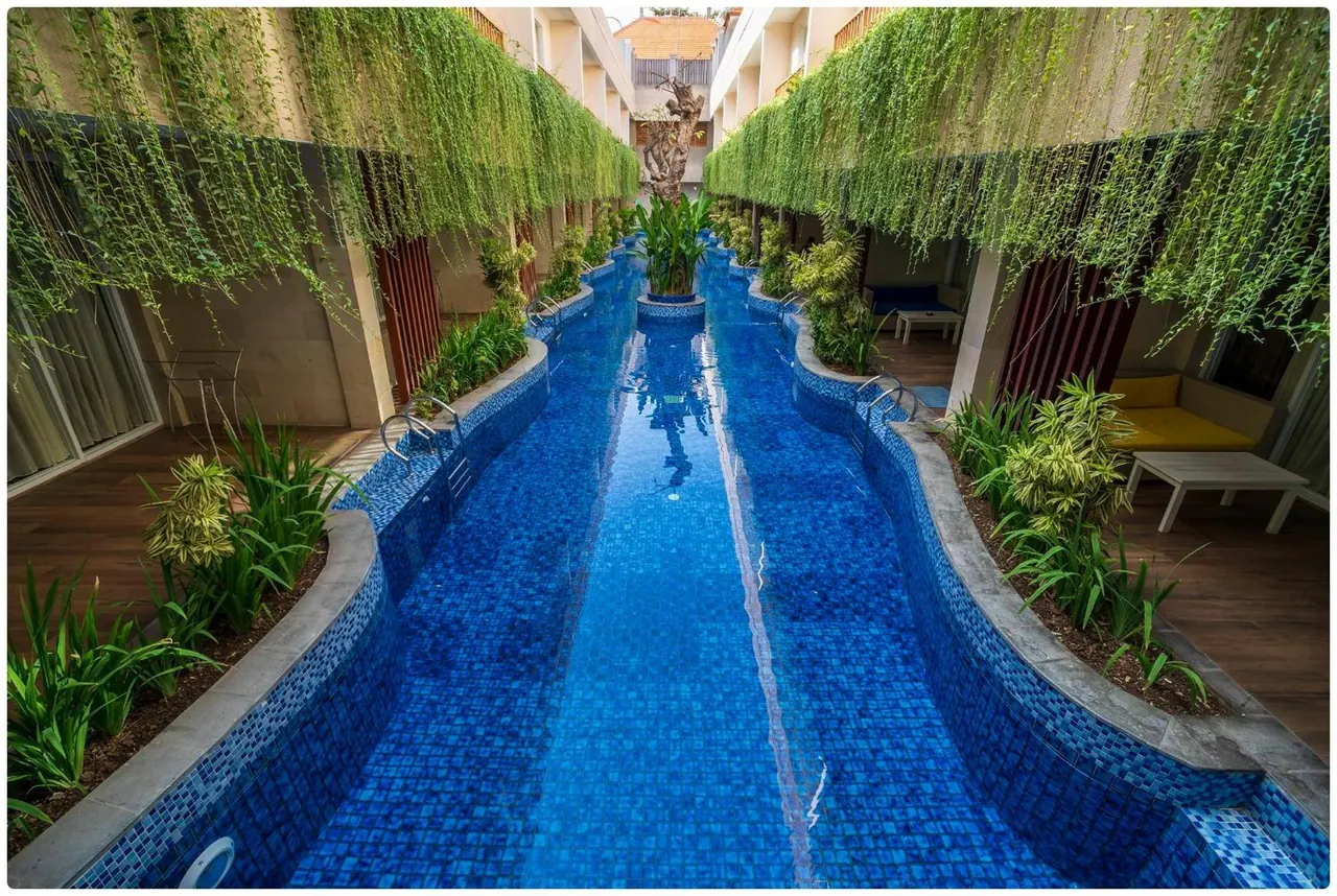 A long narrow swimming pool with blue mosaic tiles runs between two rows of rooms, with hanging vines overhead and planted beds along its edges.