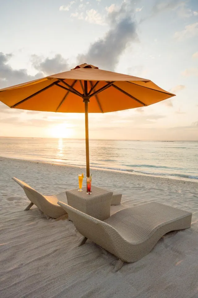 Two wicker loungers, a side table with two drinks and a yellow parasol stand on a sandy beach facing the sea at sunset.