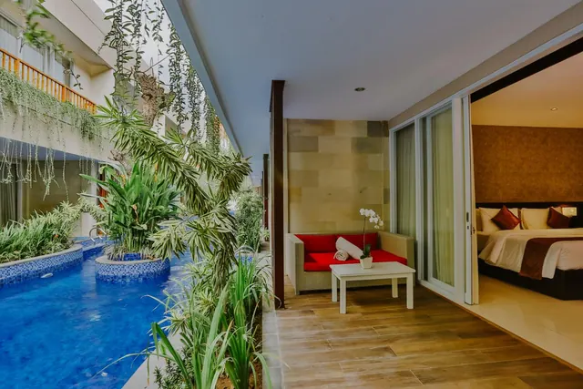 A covered terrace with a red-cushioned wicker sofa and white coffee table sits beside an open sliding door to a bedroom with a double bed, with a pool and tropical plants alongside.