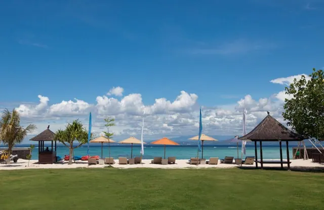 Sun loungers, thatched gazebos and parasols sit on a sandy beach in front of a calm blue sea under a partly cloudy sky.