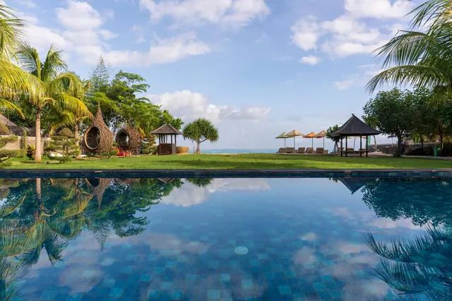 A tiled pool in the foreground reflects the sky, with a lawn, woven bamboo pavilions, sun loungers under umbrellas and the sea in the distance.