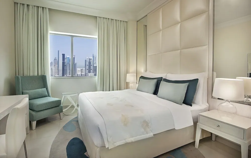 A bedroom with a made king bed, a teal armchair, a white desk and a window showing a city skyline.