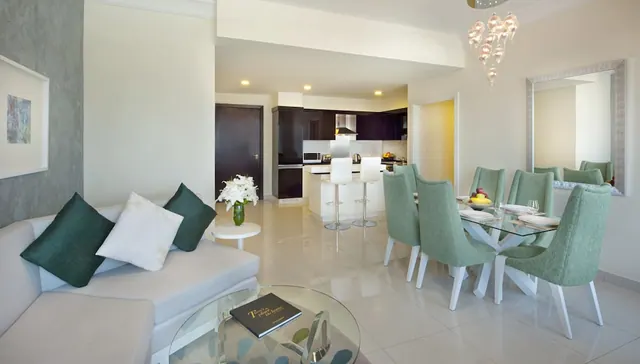 A bright living and dining area with a white sofa, green cushions, a glass dining table with green chairs, and an open kitchen with bar stools.