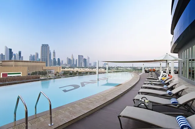 A rooftop swimming pool with sun loungers and shaded seating, with a city skyline of tall towers in the background.