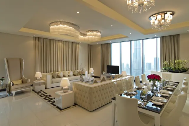 A living and dining area with cream sofas, crystal chandeliers, a dining table set for a meal, and floor-to-ceiling windows showing the Burj Khalifa and city skyline.