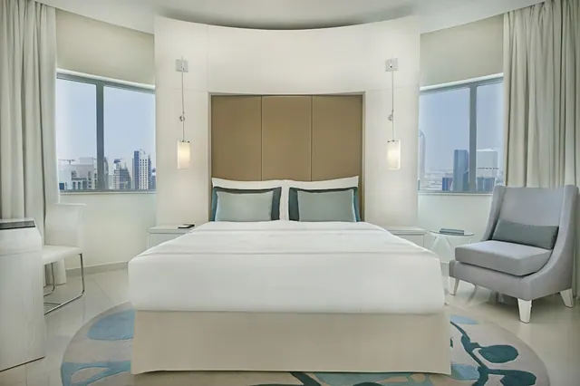 A bedroom with a made king bed, a curved headboard wall, a grey armchair, and two windows overlooking city towers.