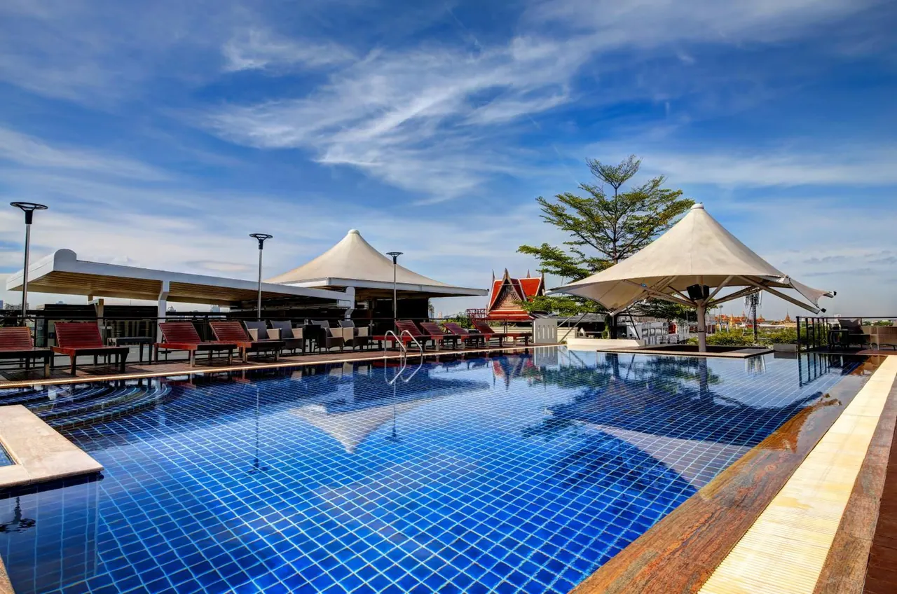 A rooftop swimming pool with blue mosaic tiles, red sun loungers and white shade canopies under a blue sky.