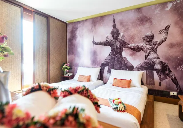 A bedroom with two beds, orange cushions and flower garlands on the bedding, in front of a large mural of two Thai giant figures.