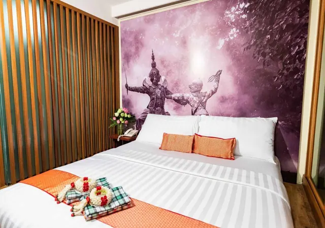 A double bed with white striped linen, an orange runner and orange cushions sits in front of a purple wall mural of two Thai mythical figures.