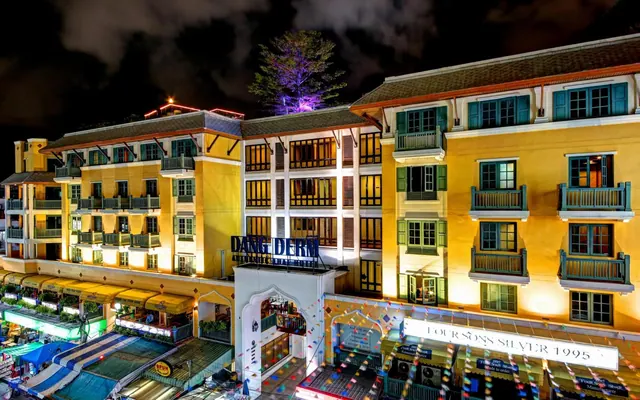 Night exterior of a multi-storey yellow hotel building with illuminated balconies, a Dang Derm sign on the upper facade and a Four Sons Silver 1995 sign over the street-level shopfronts.