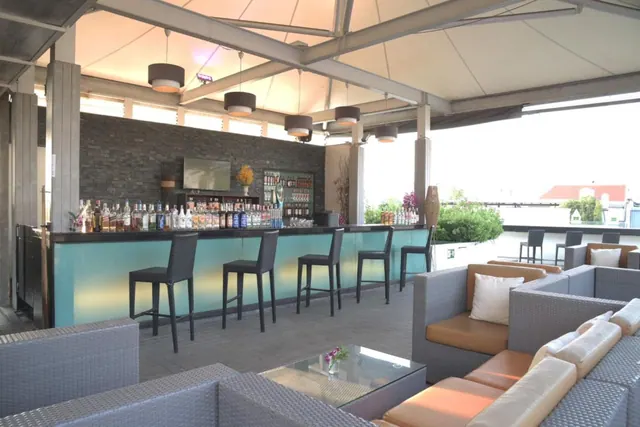 A rooftop bar with a teal-fronted counter, bar stools, shelves of bottles and wicker lounge seating under a canopy.