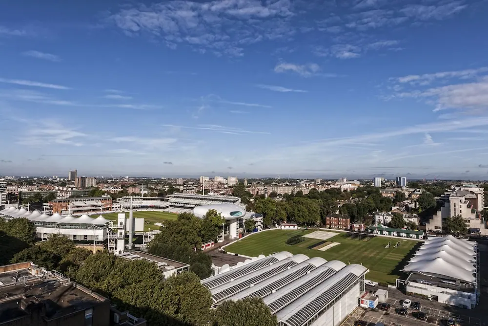 Aerial view from an upper floor across Lord's Cricket Ground and surrounding rooftops towards the London skyline under a blue sky with scattered clouds.