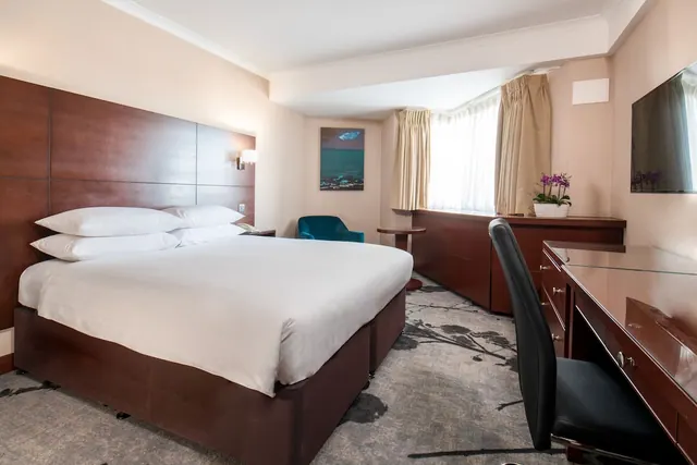 A hotel bedroom with a double bed, a teal armchair, a wooden desk with a black chair, and a wall-mounted television.