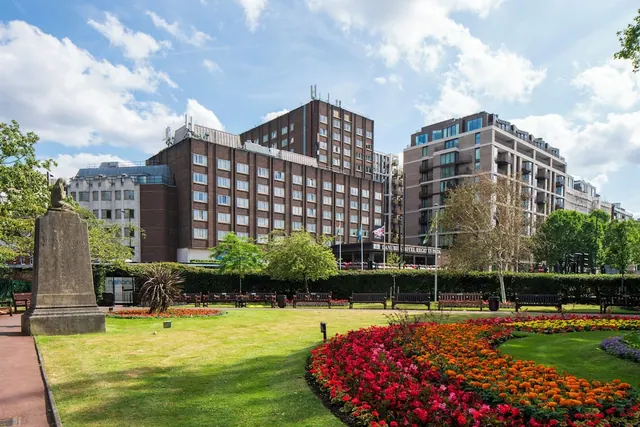 A flower-filled garden with a stone statue and park benches, with a large brick hotel building and a modern apartment block behind it under a partly cloudy sky.