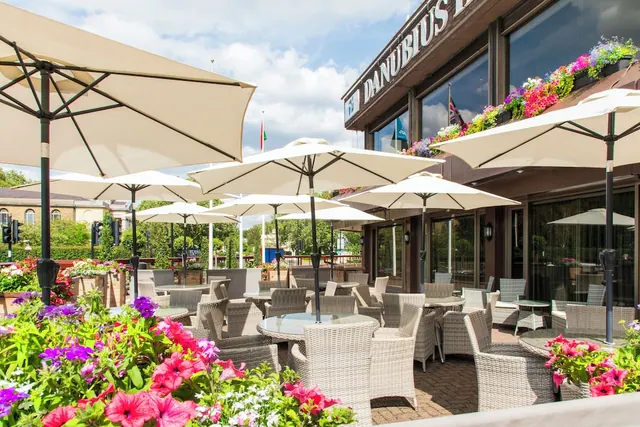 An outdoor terrace with cream parasols, wicker chairs and glass-topped tables sits beside a building with a Danubius Hotel sign and flower planters.