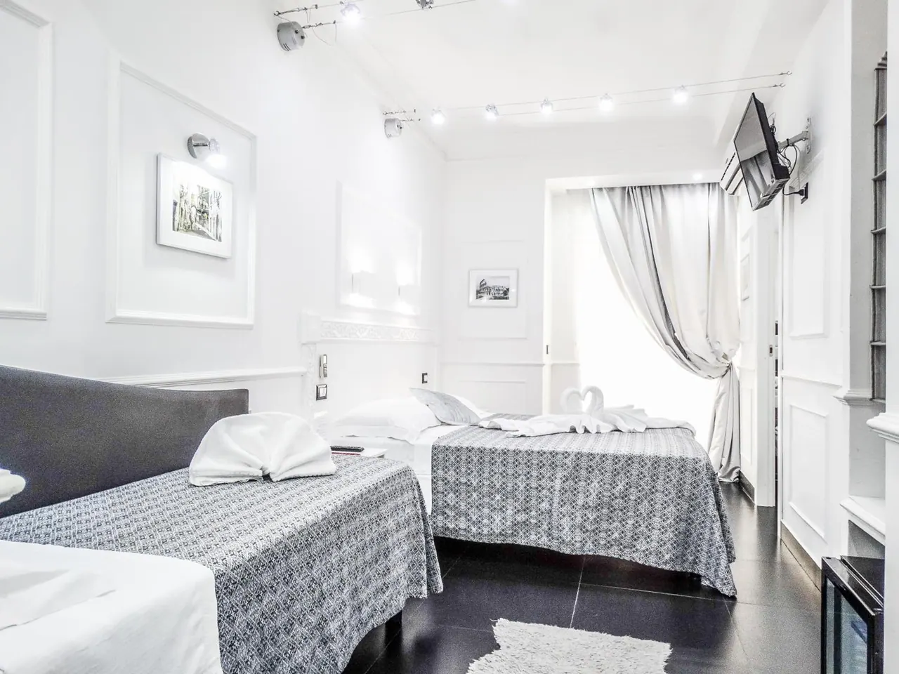 A bedroom with two beds with grey patterned covers, white towel swans on the beds, a wall-mounted television, and curtains beside a bright window.