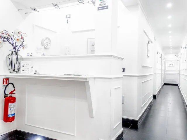 A white reception desk with a wall clock, a vase of artificial orchids and a red fire extinguisher, with a long corridor with dark floors running beyond it.