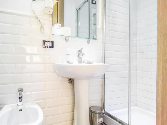 A white tiled bathroom with a pedestal sink, a mirror above it with a glass shelf, a bidet on the left, a glass-enclosed walk-in shower and a stainless steel bin.