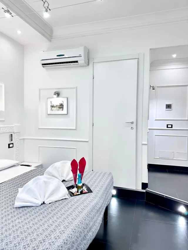A white bedroom with a grey patterned bed, two white towel swans, a tray with red napkins and a champagne bottle, a wall-mounted air conditioner and a white door.