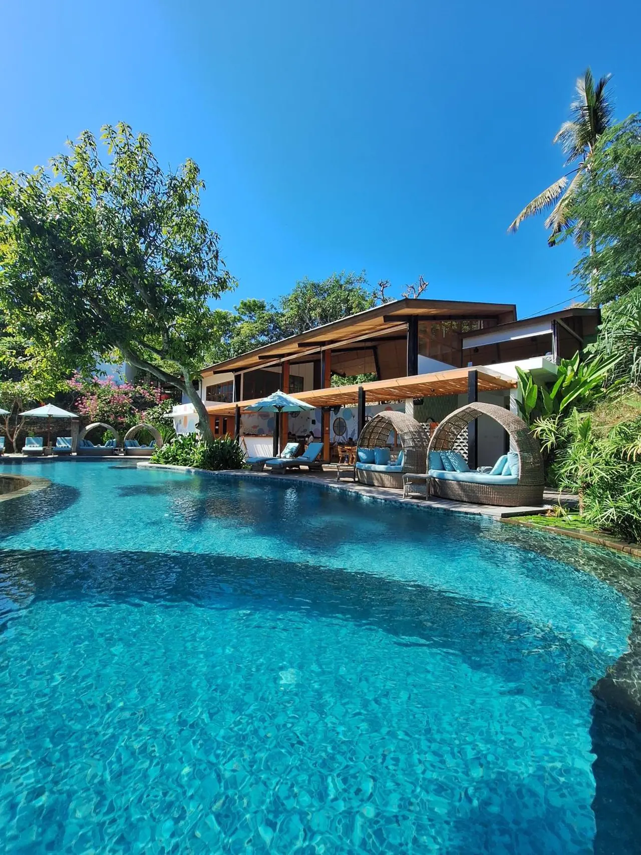 A large turquoise swimming pool with sun loungers and canopied wicker daybeds beside a wooden-roofed building and tropical trees under a clear blue sky.