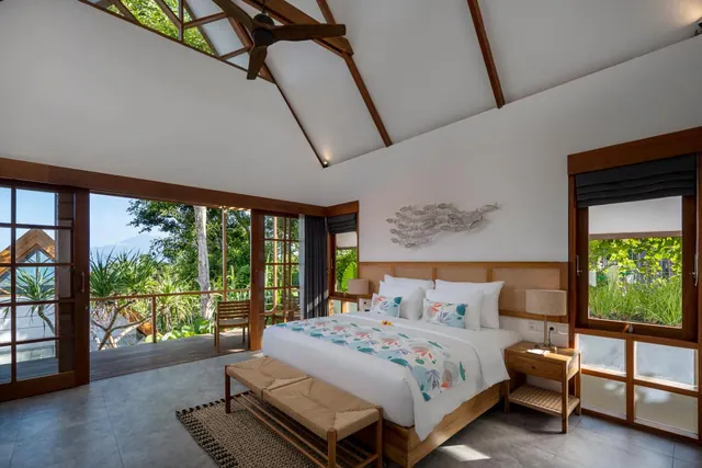 A bedroom with a king bed, a vaulted timber ceiling and a ceiling fan, with glass doors opening onto a deck overlooking trees and greenery.
