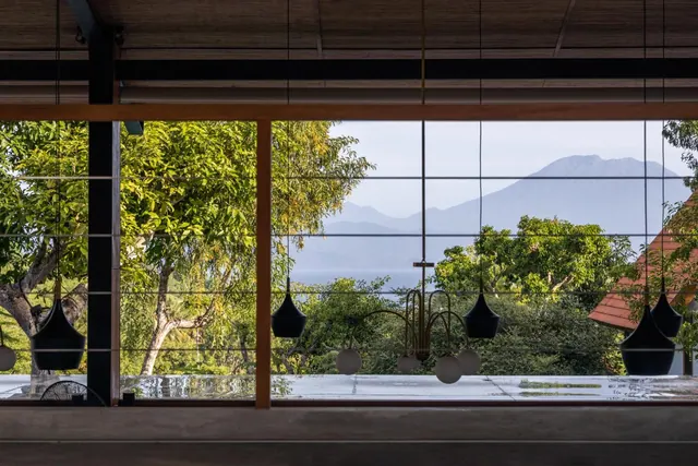 A large window frames a view of green trees, a strip of blue sea and a hazy volcanic mountain, with black pendant lights hanging in the foreground.