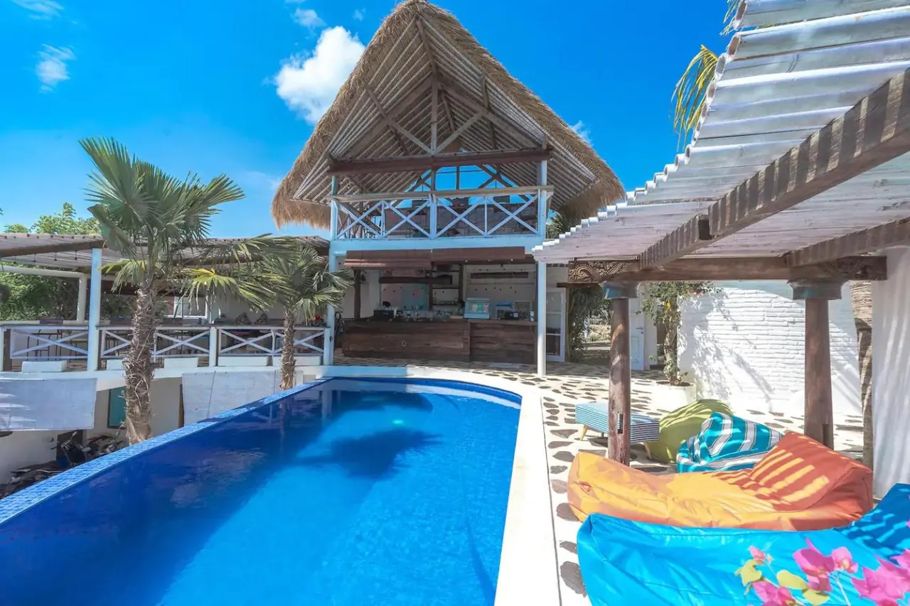 A long blue swimming pool sits in front of a thatched-roof building with an open bar area, with bean bag cushions and a wooden pergola to the right.