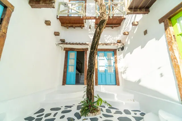 A whitewashed courtyard with blue doors, a tree trunk rising beside a planted bed on stone pavers, and a wooden balcony above.