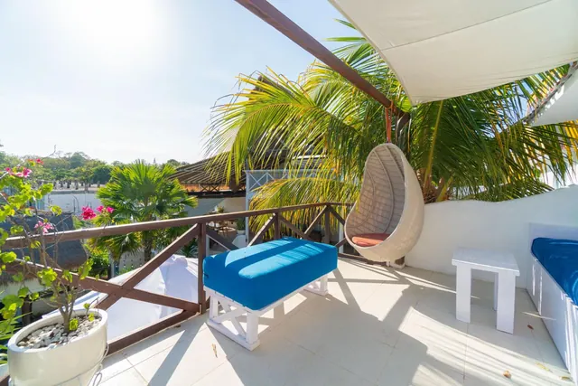 A tiled balcony with a hanging wicker egg chair, a blue cushioned bench, a white side table, a wooden railing and palm trees beyond.
