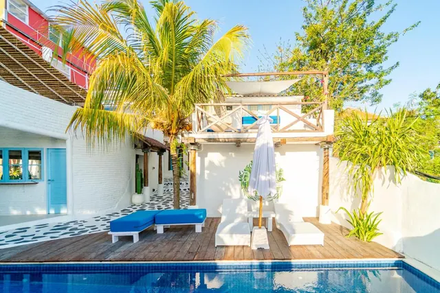 A small pool beside a wooden deck with blue daybeds and white loungers, a closed white umbrella, palm trees and a white building with an upper balcony behind.