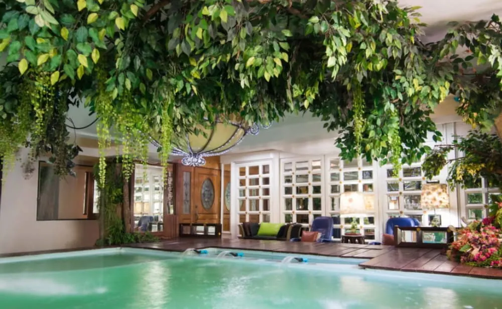 An indoor pool sits beneath hanging green plants, with a wooden deck, sofas and armchairs beside it.