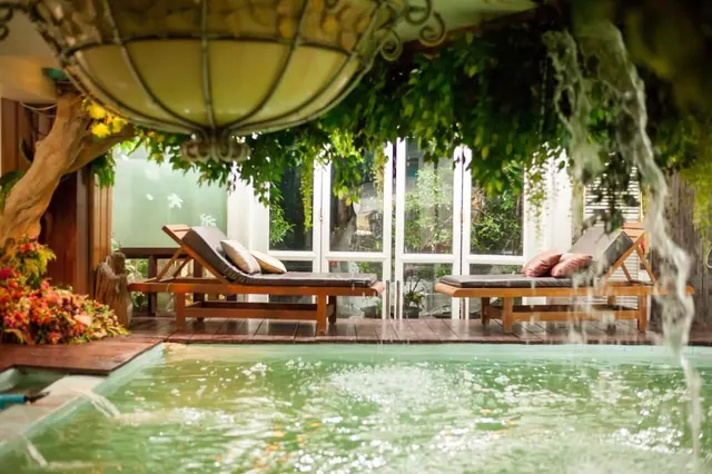 An indoor pool with a water feature cascading into it, beside two wooden sun loungers with cushions, under a hanging lantern and green plants, with glass doors onto a garden behind.