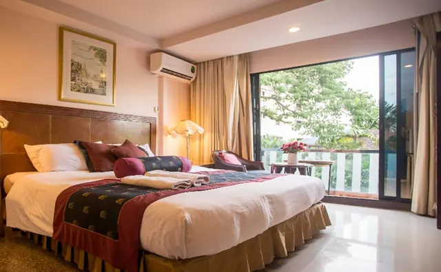 A bedroom with a made king bed, a wall-mounted air conditioner, and a glass sliding door opening onto a balcony with trees outside.