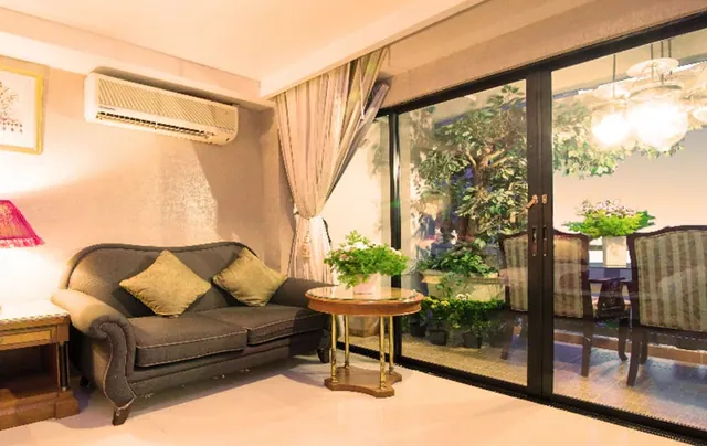 A living room with a grey sofa and two cushions, a wall-mounted air conditioner, and sliding glass doors opening onto a balcony with potted plants and chairs.