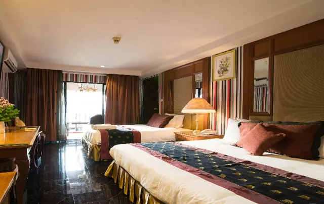 A hotel bedroom with a large double bed in front and a second bed behind it, a bedside lamp, a wood-panelled headboard wall, and a glass door to the balcony at the back.