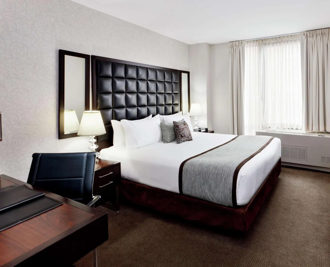 A bedroom with a king bed, a tufted black leather headboard, a wooden desk with a leather chair, and a large window with sheer curtains.