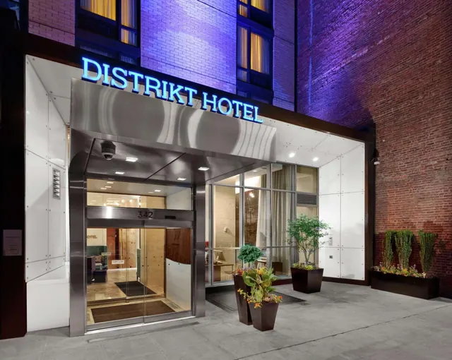 Night view of the Distrikt Hotel street entrance with illuminated blue signage, glass doors, and potted plants on the sidewalk.