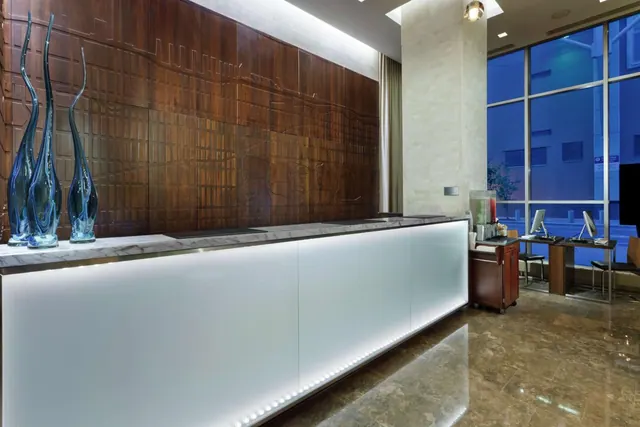 A hotel lobby with a backlit white reception desk, a carved wooden wall panel, blue glass sculptures, and a computer workstation beside tall windows.