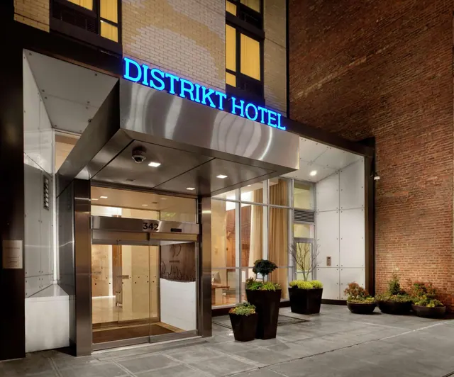 Entrance of the Distrikt Hotel at dusk, with illuminated blue signage, a glass door marked 342, and potted plants along the sidewalk beside a brick wall.