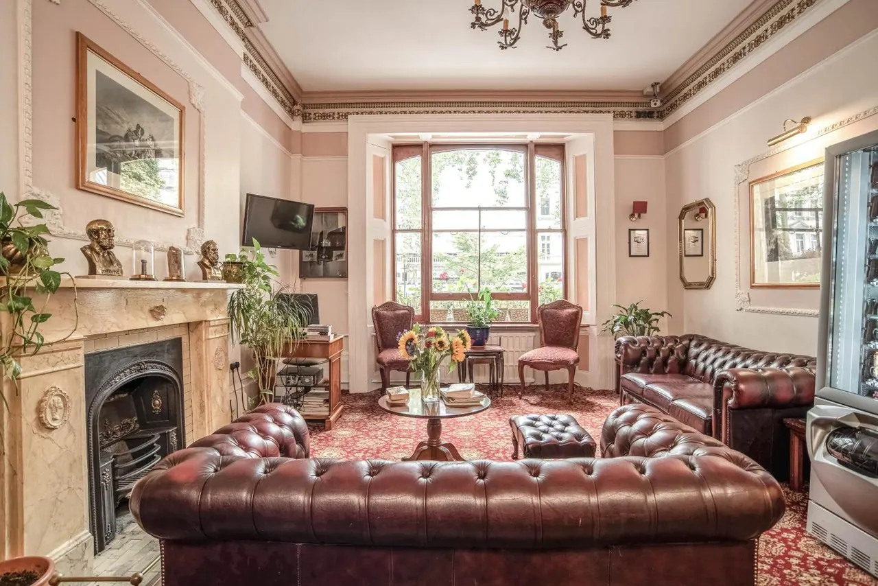 A lounge with tufted brown leather chesterfield sofas, a marble fireplace and a tall bay window looking out onto trees and a street.