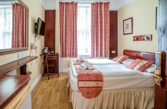 A double bed with red and cream bedding and folded towels sits in a bedroom with red curtains, a wall-mounted television and a small wooden desk holding a kettle.