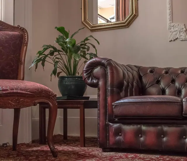 A leather chesterfield sofa, a carved wooden chair, a potted plant on a side table and a gold framed mirror sit in a room with a patterned rug.