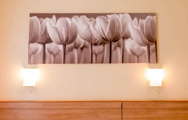 A bedroom wall with a large sepia tulip canvas print above, flanked by two lit wall lamps and a wooden surface below.