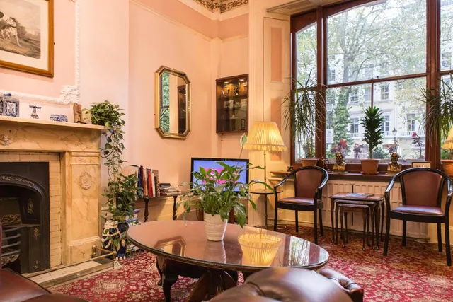 A sitting room with a marble fireplace, leather armchairs, a round glass-topped table and tall windows looking onto a tree-lined street.