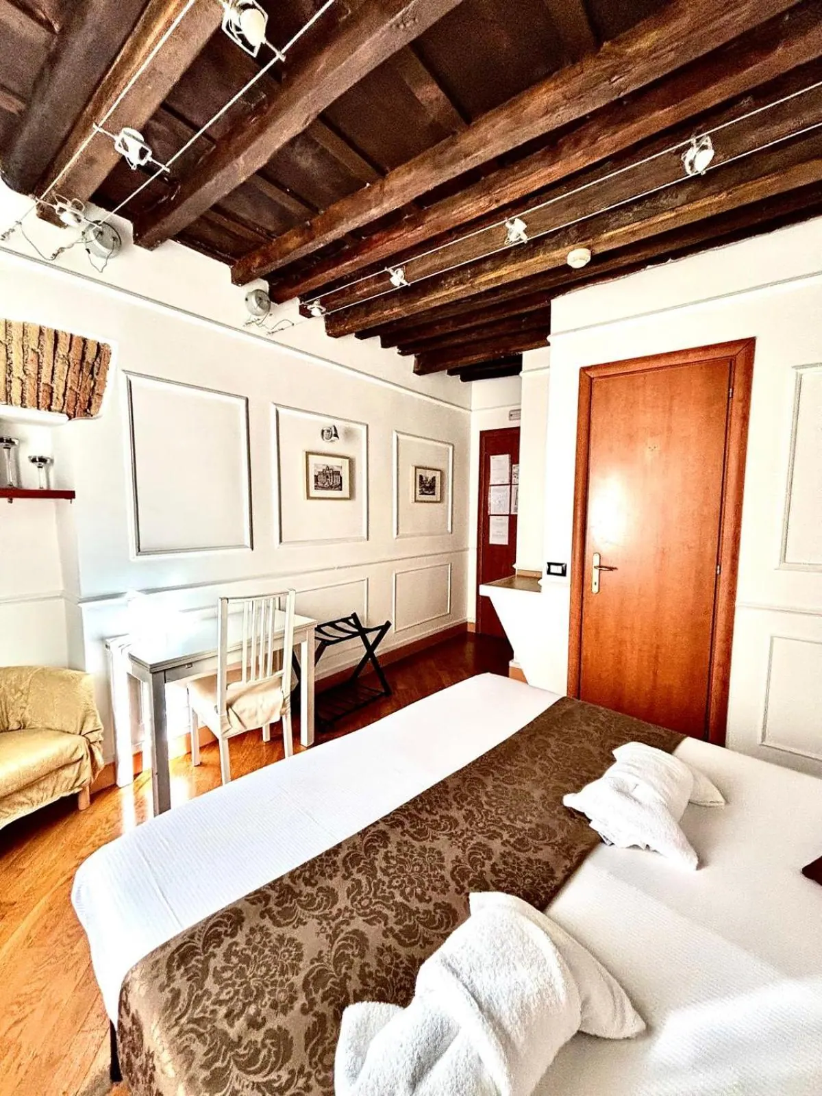 A bedroom with exposed dark wooden ceiling beams, a double bed with white linen and a brown damask runner, a small desk with a chair, and a beige armchair against the left wall.