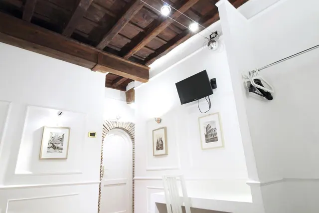 A white-walled room with exposed wooden ceiling beams, a wall-mounted flat-screen TV, framed prints and a white chair at the bottom edge of the frame.